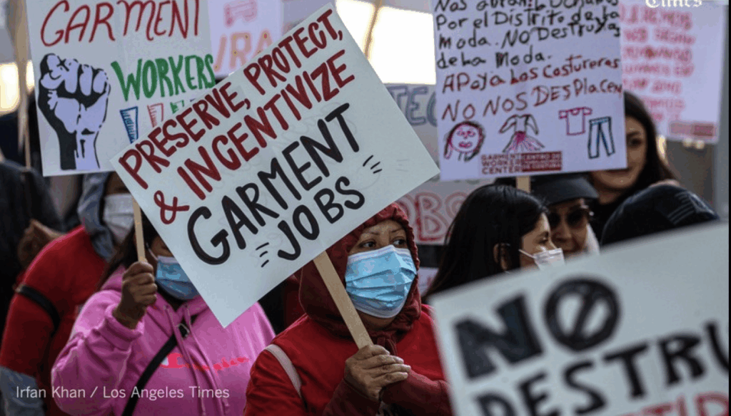 Protestors carry home made picket signs bearing slogans about protecting the LA garment district. In the center of the frame, a woman in a mask carries a sign reading "Preserve, Protect, & Incentivize Garment Jobs"